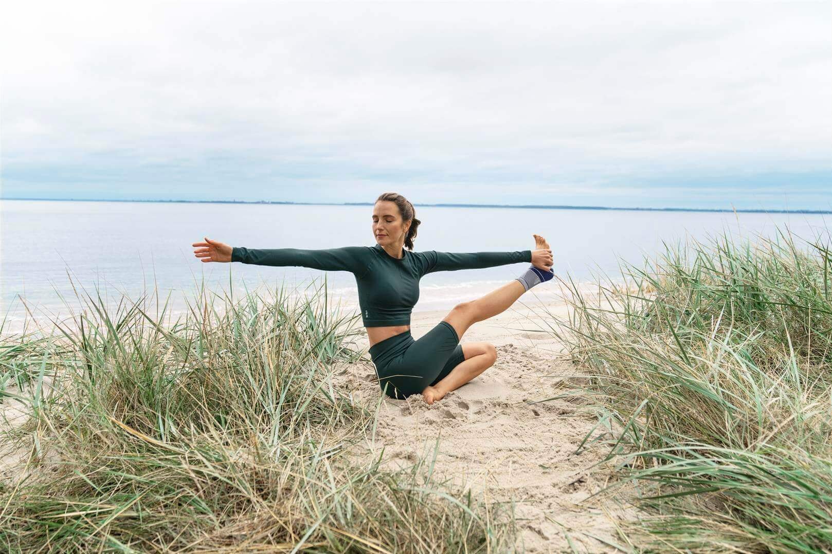 Das Bild zeigt eine Frau, die am Strand eine Yoga-Pose einnimmt. Sie trägt die MalleoTrain Sprunggelenkbandage von Bauerfeind, um Sprunggelenkschmerzen entgegenzuwirken.