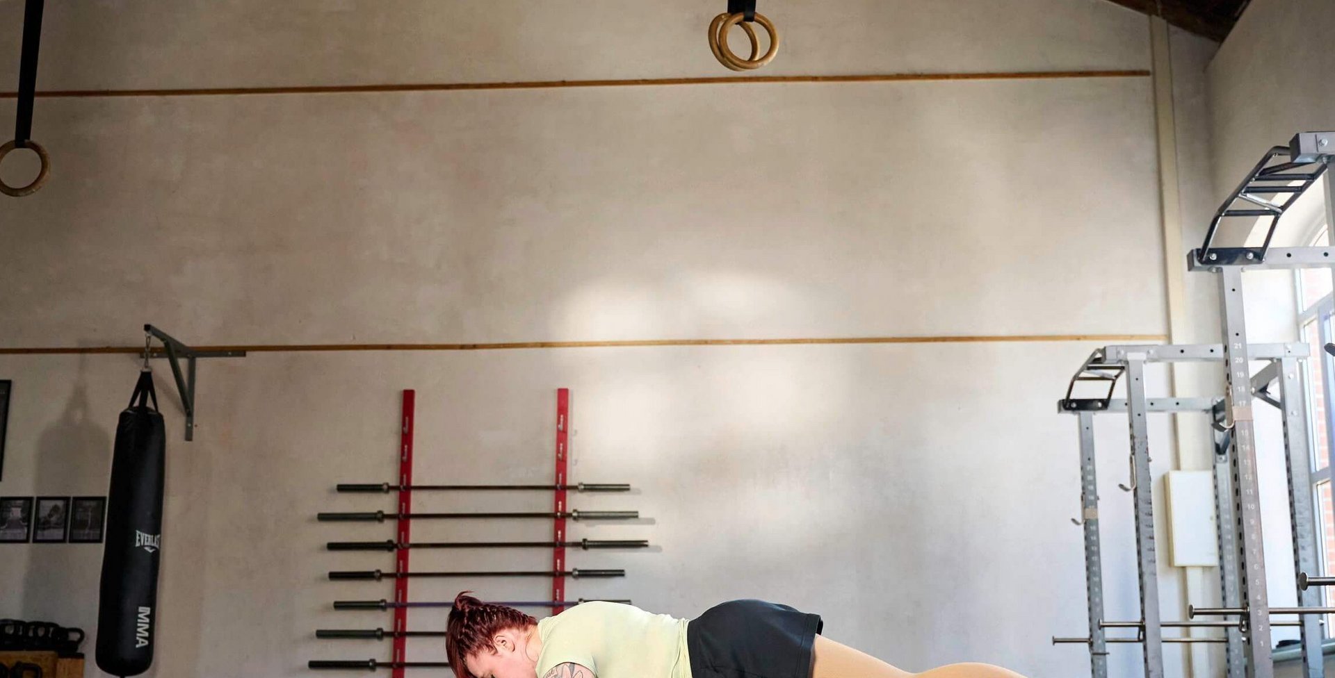Person macht eine Unterarmstütz-Übung (Plank) auf einer Fitnessmatte in einem geräumigen Trainingsraum mit hohen Decken, Hantelstangen an der Wand und Fenstern auf der rechten Seite.