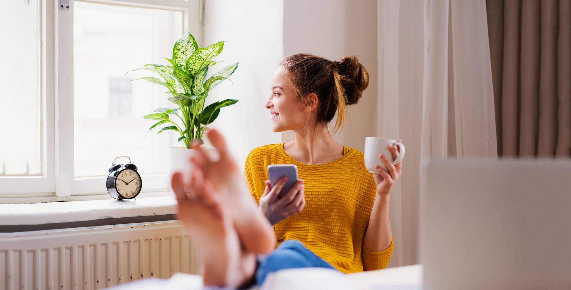 Frau am Schreibtisch Auf dem Bild sitzt eine junge Frau auf einem Stuhl und hat ihre Füße auf den Tisch vor ihr abgelegt. In der linken Hand hält sie eine Tasse; in der rechten Hand ein Smartphone. Regelmäßiges Training der Fußmuskulatur kann Schmerzen vorbeugen.