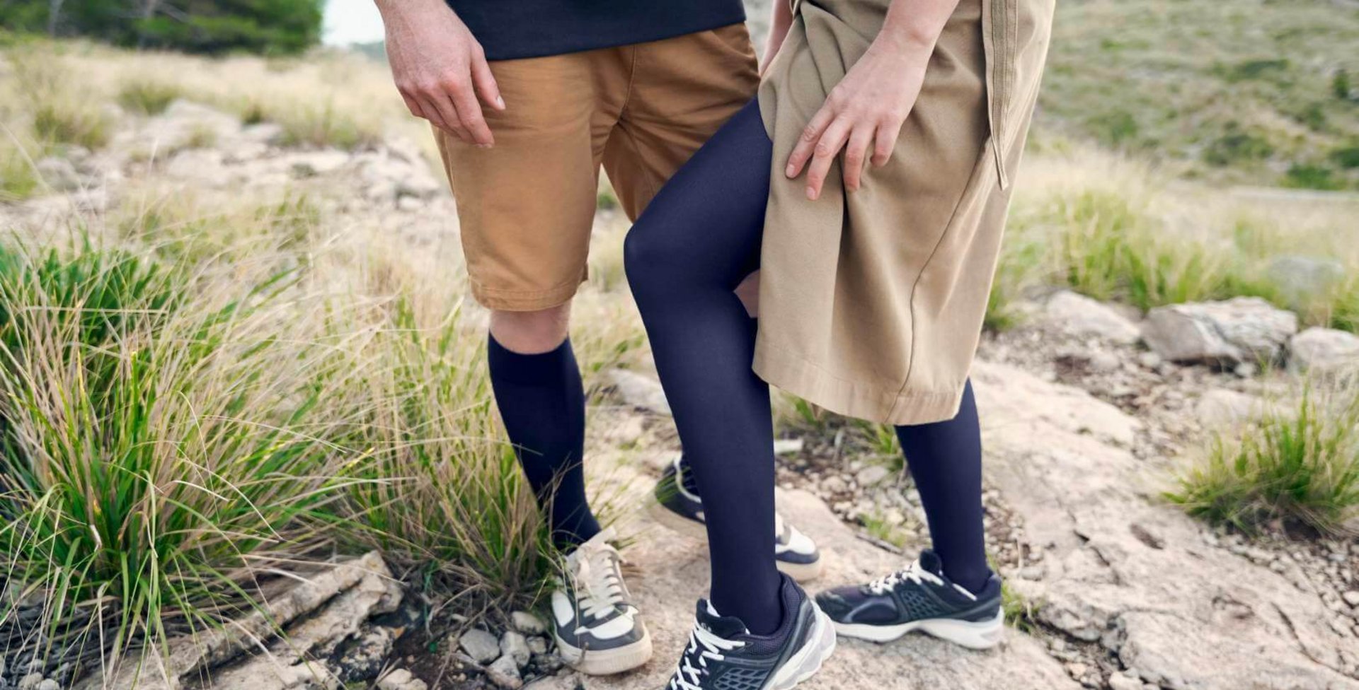 Nahaufnahme von zwei Personen in einer felsigen Landschaft, beide tragen dunkelblaue VenoTrain Kompressionsstrümpfe von Bauerfeind, kombiniert mit sportlichen Schuhen; eine Person trägt eine beigefarbene Hose, die andere einen beigen Rock.