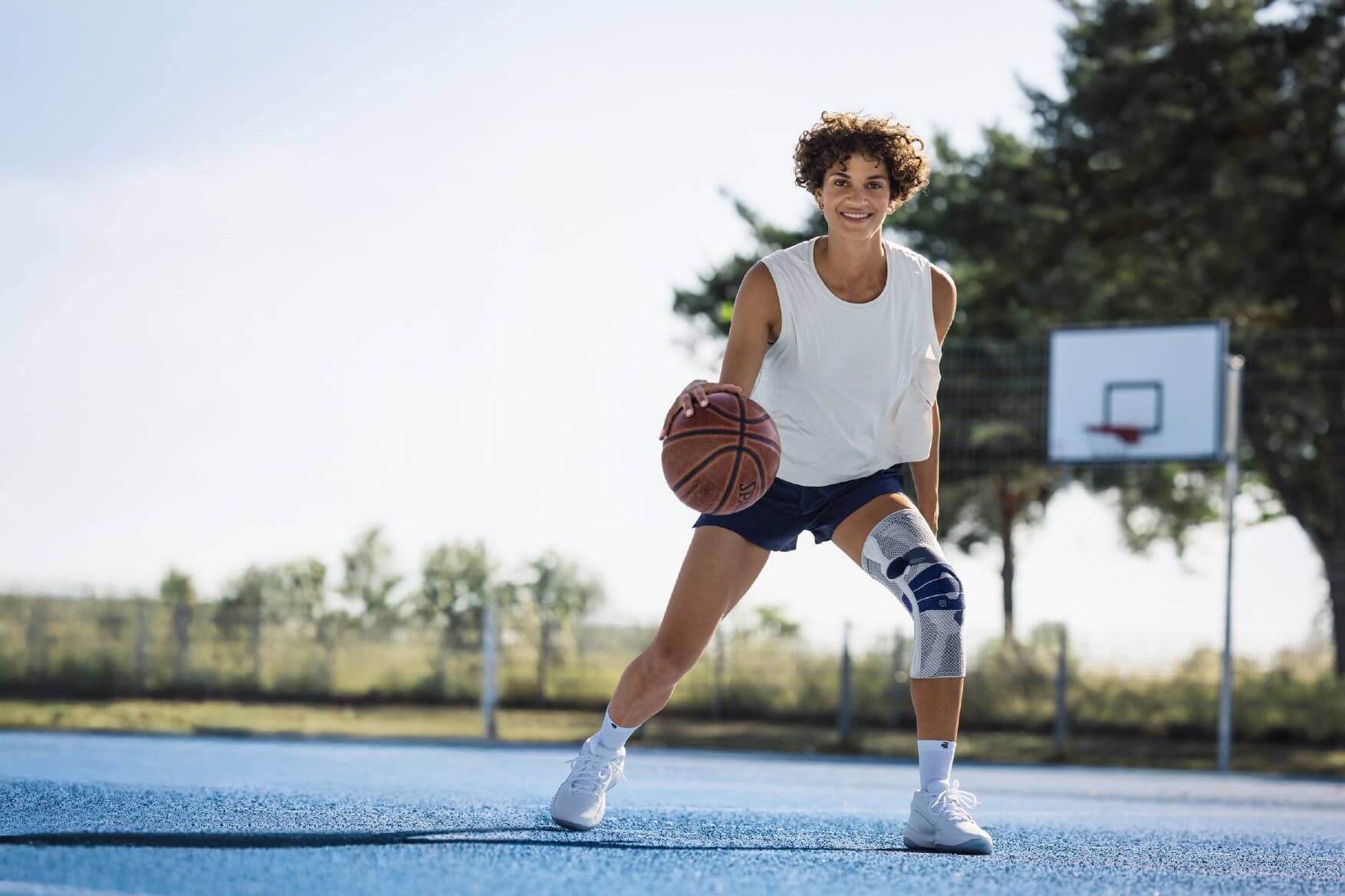 Eine Person mit weissem Trägershirt, dunklen Shorts und Kniebandage am rechten Bein dribbelt einen Basketball auf einem blauen Aussenplatz. Im Hintergrund ist ein Basketballkorb und ein Zaun sichtbar. Die Person lächelt und schaut in die Kamera.